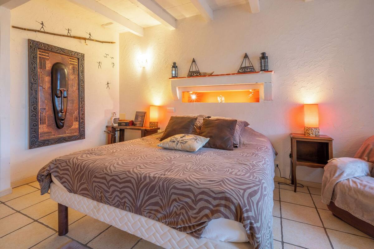 Un dormitorio con una cama grande, dos mesitas de noche, un estante en la pared con objetos decorativos y una obra de arte enmarcada sobre la cama.