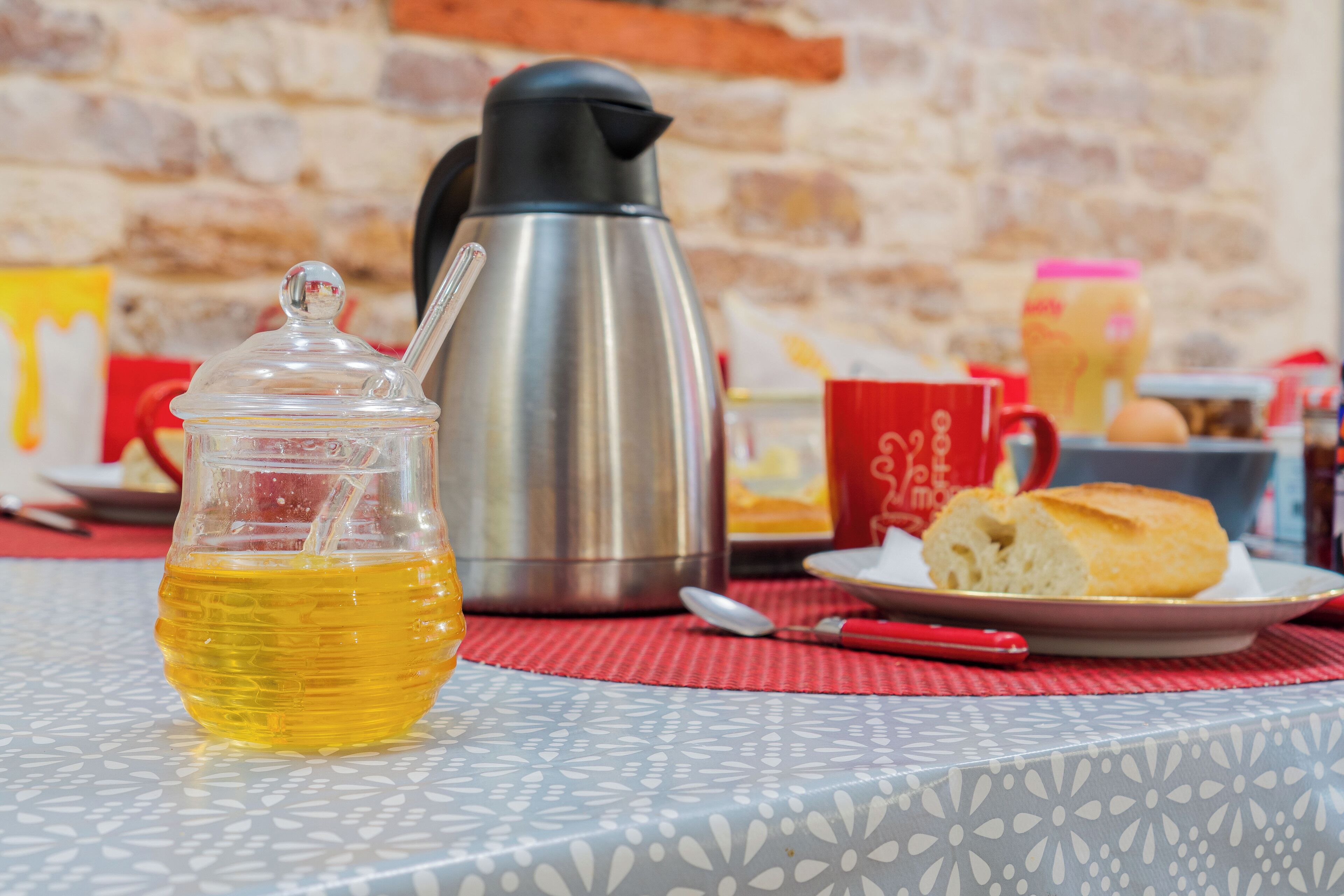 Een tafel met een glazen pot met een gele vloeistof, een roestvrijstalen waterkoker, een rode mok, een bord met een broodje en een lepel.
