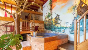 Un jacuzzi con un banco de madera, rodeado de paredes de piedra y un mural con un paisaje tropical.