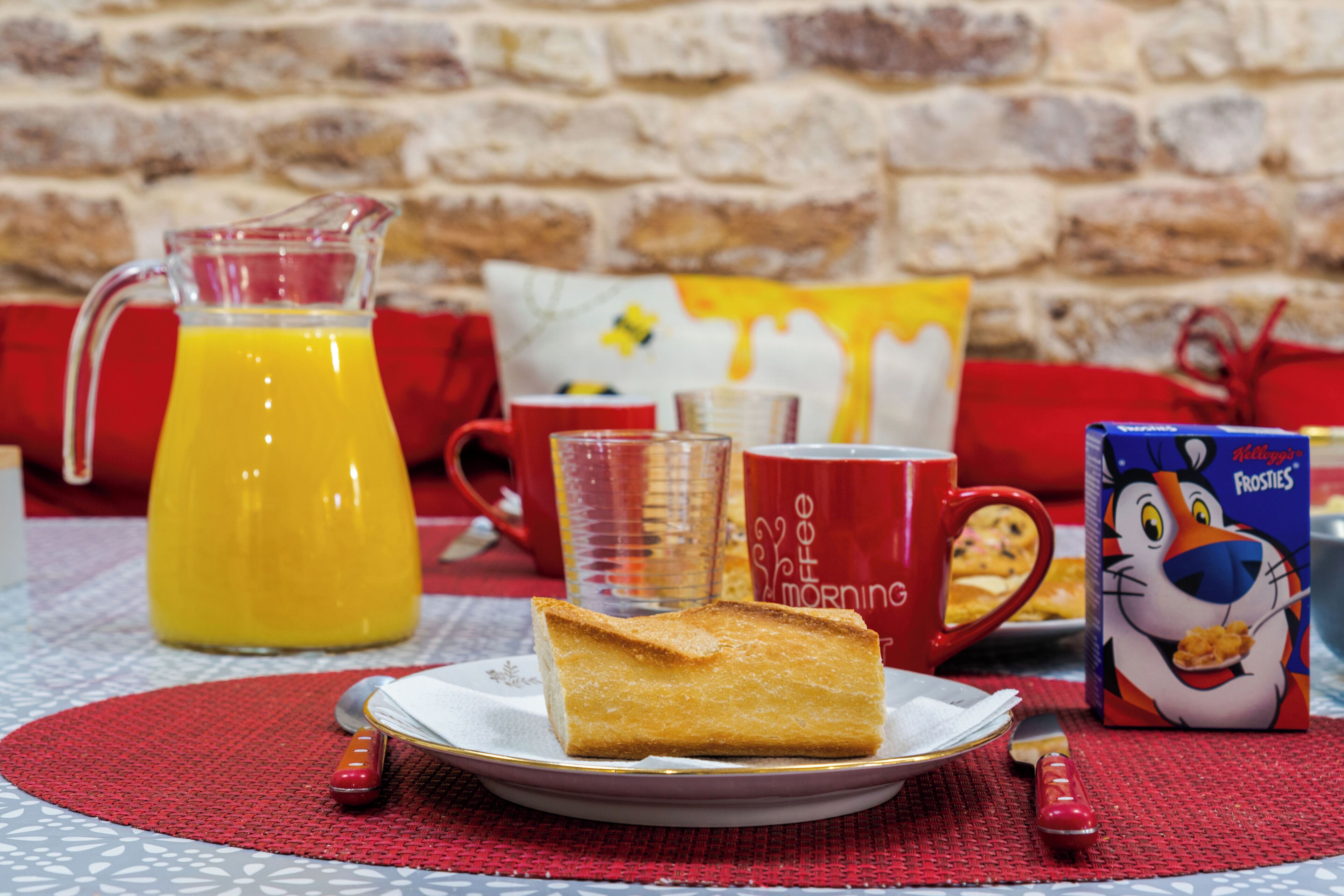 Una escena de desayuno con una jarra de jugo de naranja, un plato con una rebanada de pastel, una taza, una caja de cereal y un vaso de leche sobre una mesa con un mantel individual rojo.