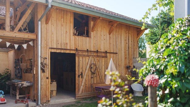 A wooden barn with a large open door, a small porch area with a table and chairs, and a garden with a potted plant. - Gîte La Schlàppe / 3 persons (HERRLISHEIM PRES COLMAR)