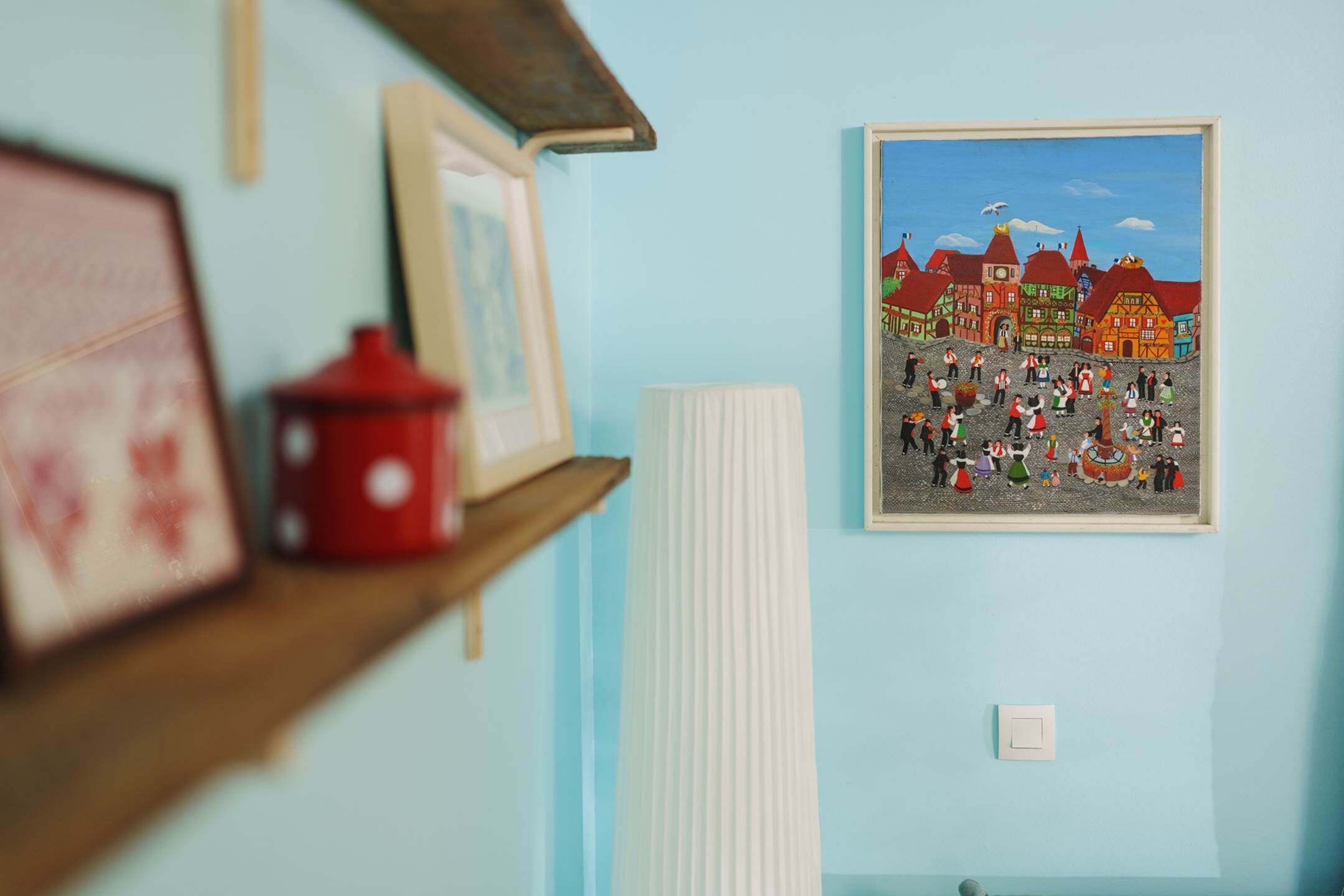 Et værelse med en lyseblå væg, et billede i ramme af et landsbymotiv, en rød potte på en hylde og en hvid radiator.