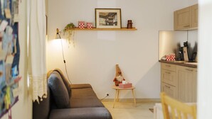 Uma sala de estar aconchegante com um sofá, uma mesinha com um ursinho de pelúcia, uma luminária e uma área de cozinha com armários e bancada.