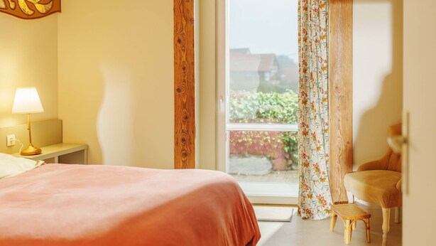 Quarto com cama, mesa de cabeceira, luminária, janela com cortinas e vista para o verde lá fora.