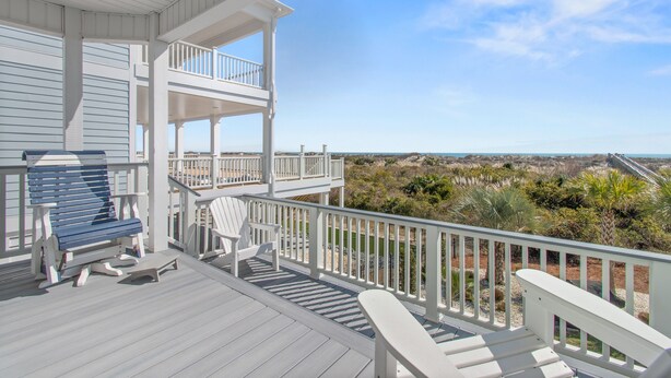 Eine Holzterrasse mit einer Bank und Stühlen, ein Balkon mit Geländern und Blick auf Strand und Dünen.