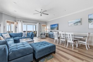 A spacious living room with a sectional sofa, a dining area with white chairs, and a ceiling fan.
