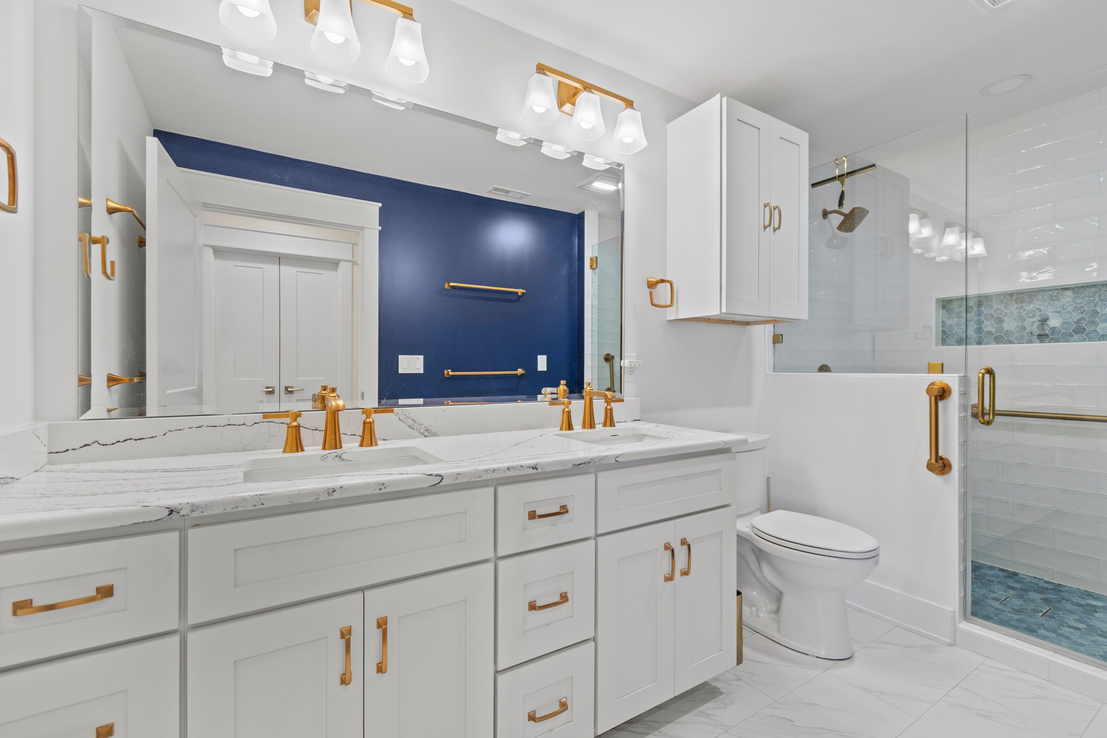 Un baño moderno con tocador blanco, detalles dorados, un espejo grande y mampara de vidrio.
