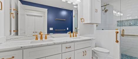 Un baño moderno con tocador blanco, detalles dorados, un espejo grande y mampara de vidrio.