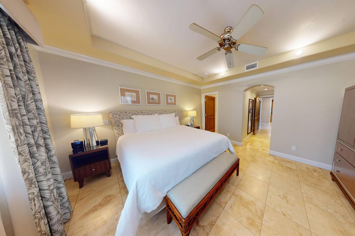 A hotel room with a large bed, a wooden bench, a wardrobe, a ceiling fan, and a nightstand with a lamp.