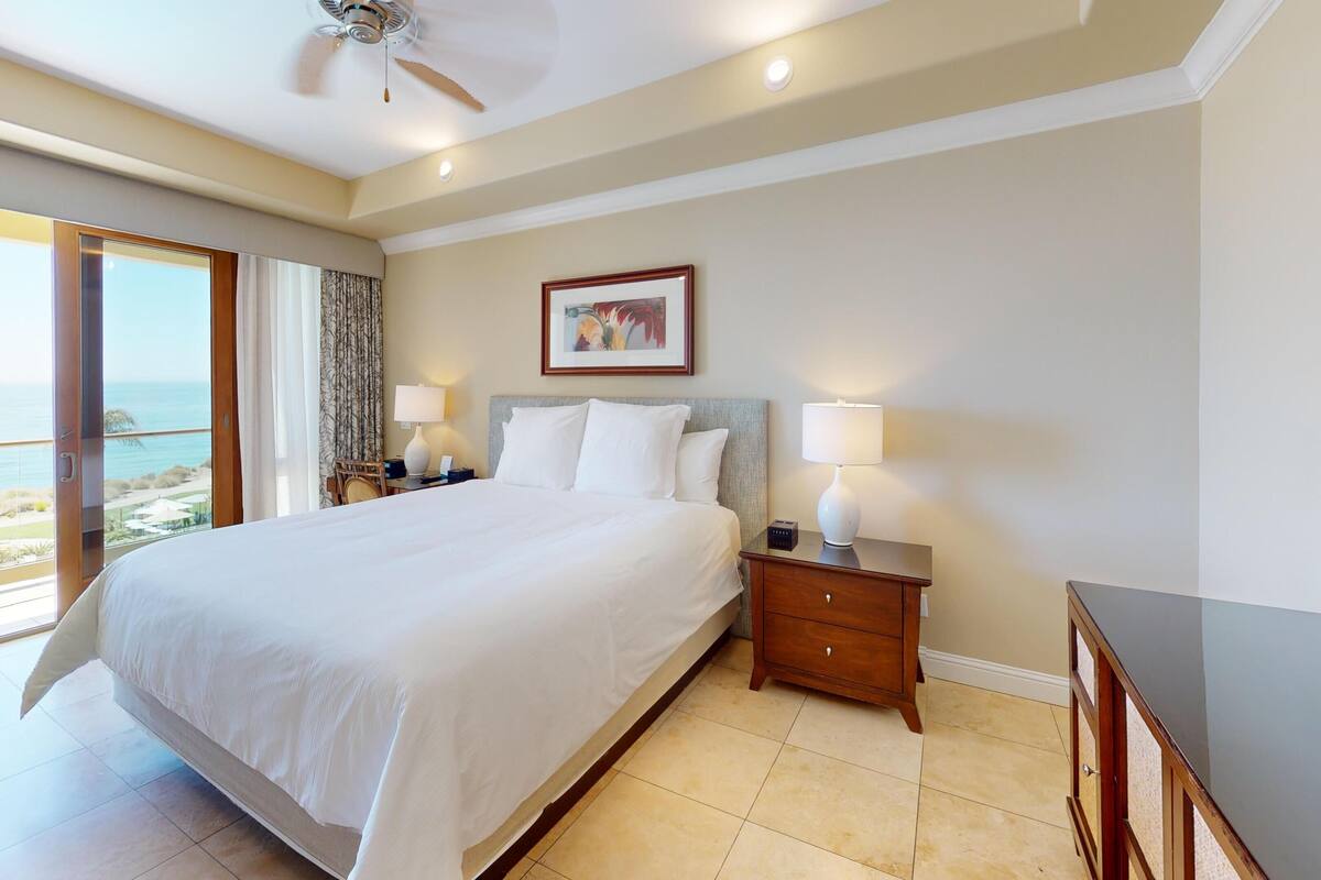 A hotel room with a large bed, bedside tables with lamps, a framed picture on the wall, and a view of the ocean.