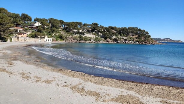 Una playa costera con casas en una colina, cielo azul despejado y mar tranquilo.