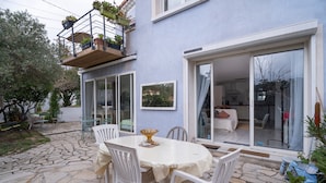 Un patio con una mesa redonda y sillas blancas, un balcón con plantas en macetas y un ventanal corredero que da a un espacio interior.