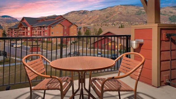 A balcony with a round table and two chairs, overlooking a residential area with red buildings and a mountainous backdrop. - Spacious  2-bedroom condo in beautiful Steamboat Springs  CO. (Steamboat Springs)