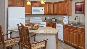 A kitchen with wooden cabinets, a white refrigerator, a microwave, a stove, a coffee maker, and a sink. There are two chairs with patterned cushions and a wooden island with a bowl. - Spacious  2-bedroom condo in beautiful Steamboat Springs  CO. (Steamboat Springs)