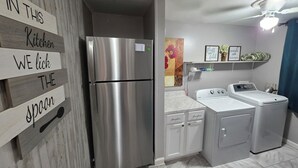 A compact kitchen with a stainless steel refrigerator, a washing machine, and a dryer. There is a wooden sign on the wall and a small shelf with decorative items. - Bluff City Studio: B (Millington)