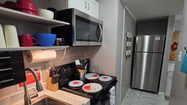 A modern kitchen with a stainless steel sink, a microwave, and a stove. There are shelves above the stove holding dishes and a cabinet with a microwave. - Bluff City Studio: B (Millington)
