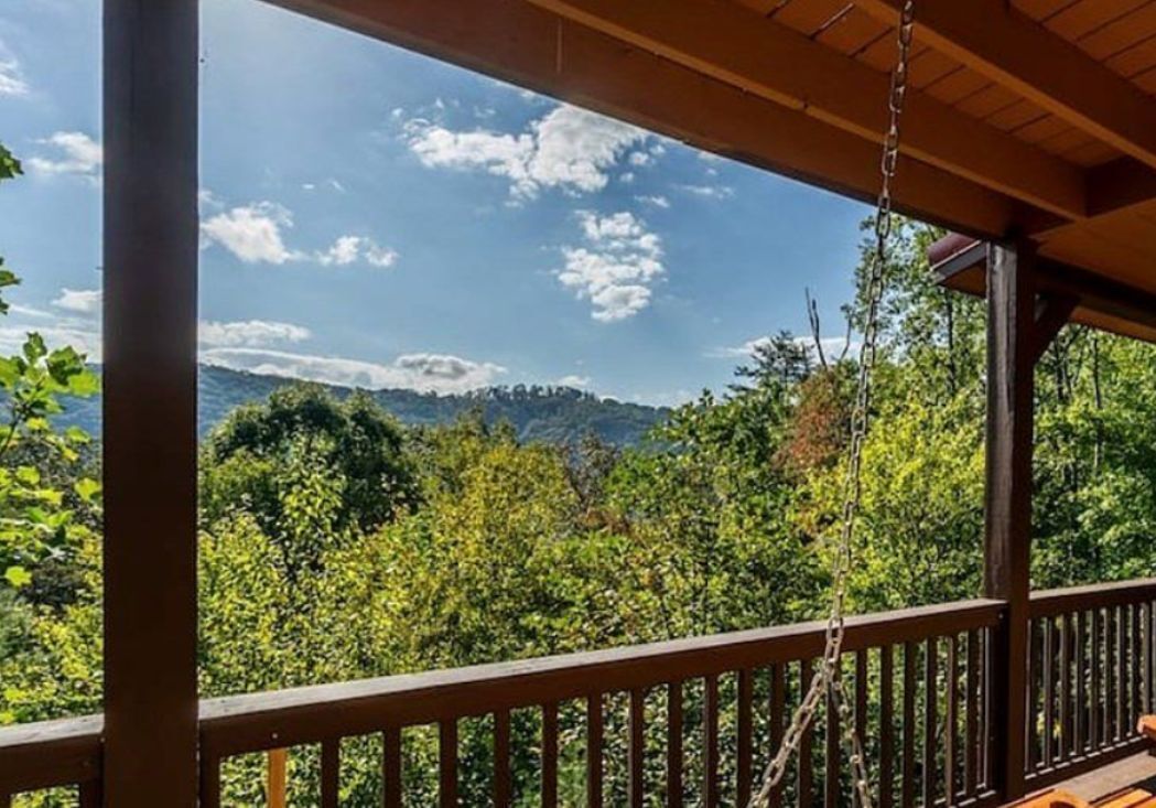 A wooden deck with a swing, overlooking a forested landscape with hills in the distance.