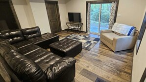 A living room with a leather sofa, a beige armchair, a TV on a stand, and a sliding glass door leading to a garden. - BEAUTY AND PRIVACY LIKE NO OTHER!   WALK TO LAKE.  CLOSE TO EVERYTHING POCONOS! (Jim Thorpe)