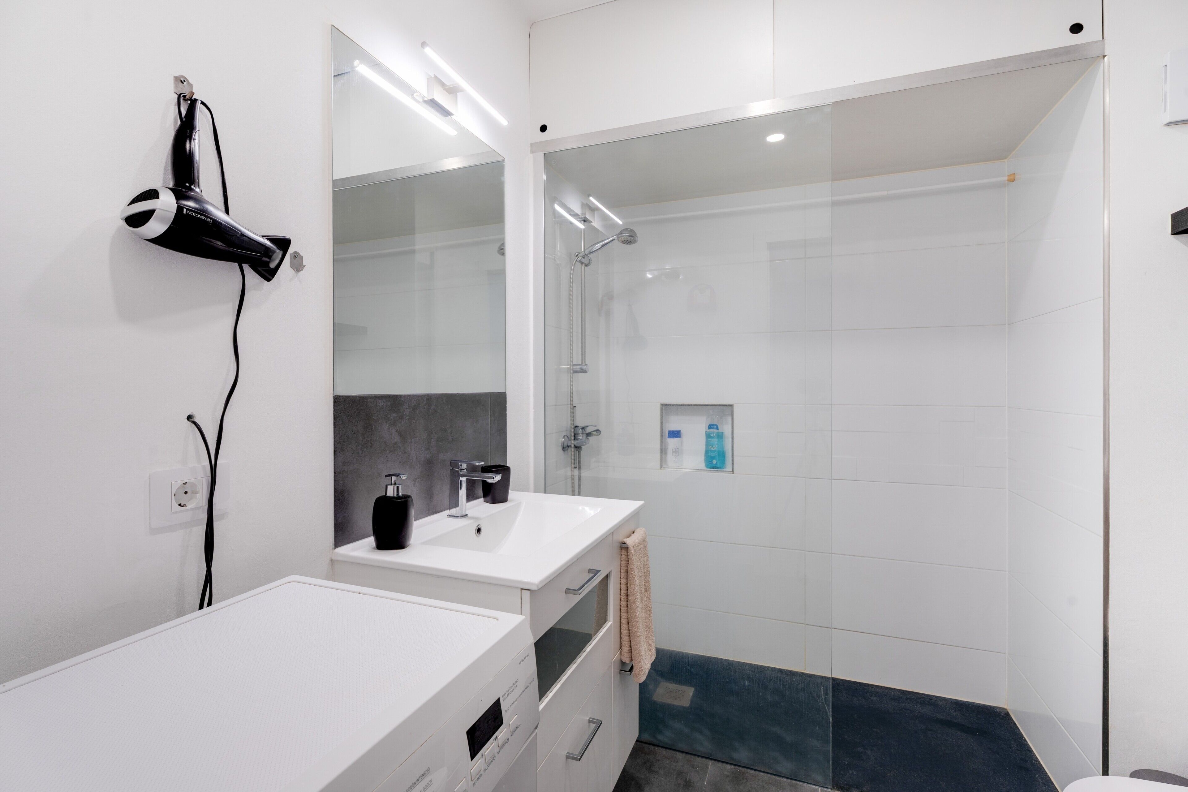 Een moderne badkamer met een wit badkamermeubel, een douchecabine van glas en een aan de muur bevestigde haardroger.
