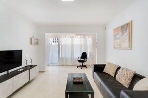 Ein Wohnzimmer mit Fernseher, einem Glastisch, einem schwarzen Ledersofa, einem schwarzen Bürostuhl und einer Glastür, die auf einen Balkon führt.