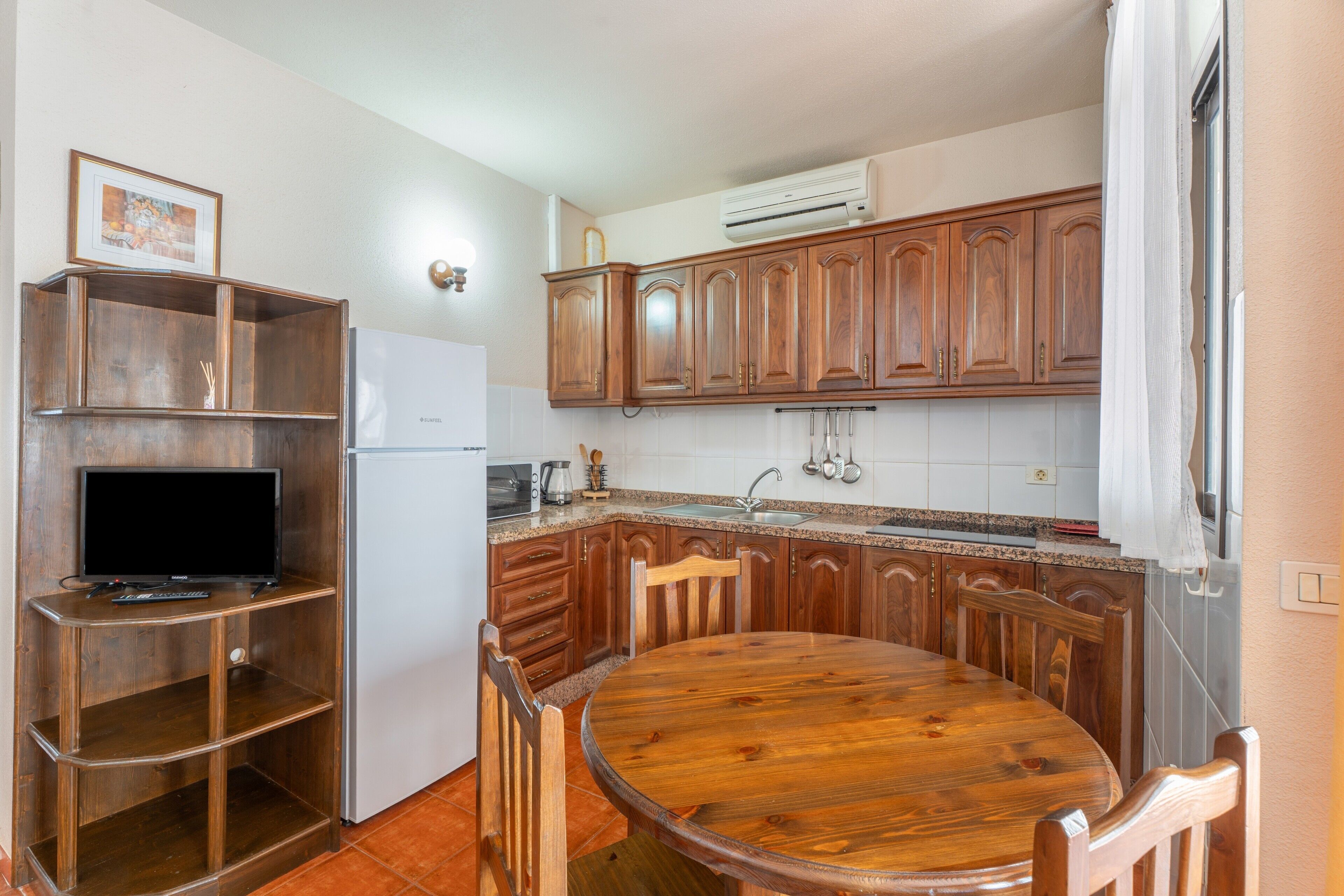 A kitchen with wooden cabinets, a built-in TV shelf, a dining table with chairs, and a refrigerator.