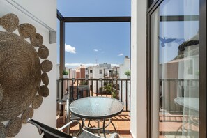 Een balkon met een glazen tafel en stoelen, uitzicht op omliggende gebouwen en een decoratieve muur.