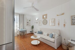 Een woonkamer met een witte bank, een ronde salontafel, een eethoek met een tafel en stoelen, en een plafondventilator.