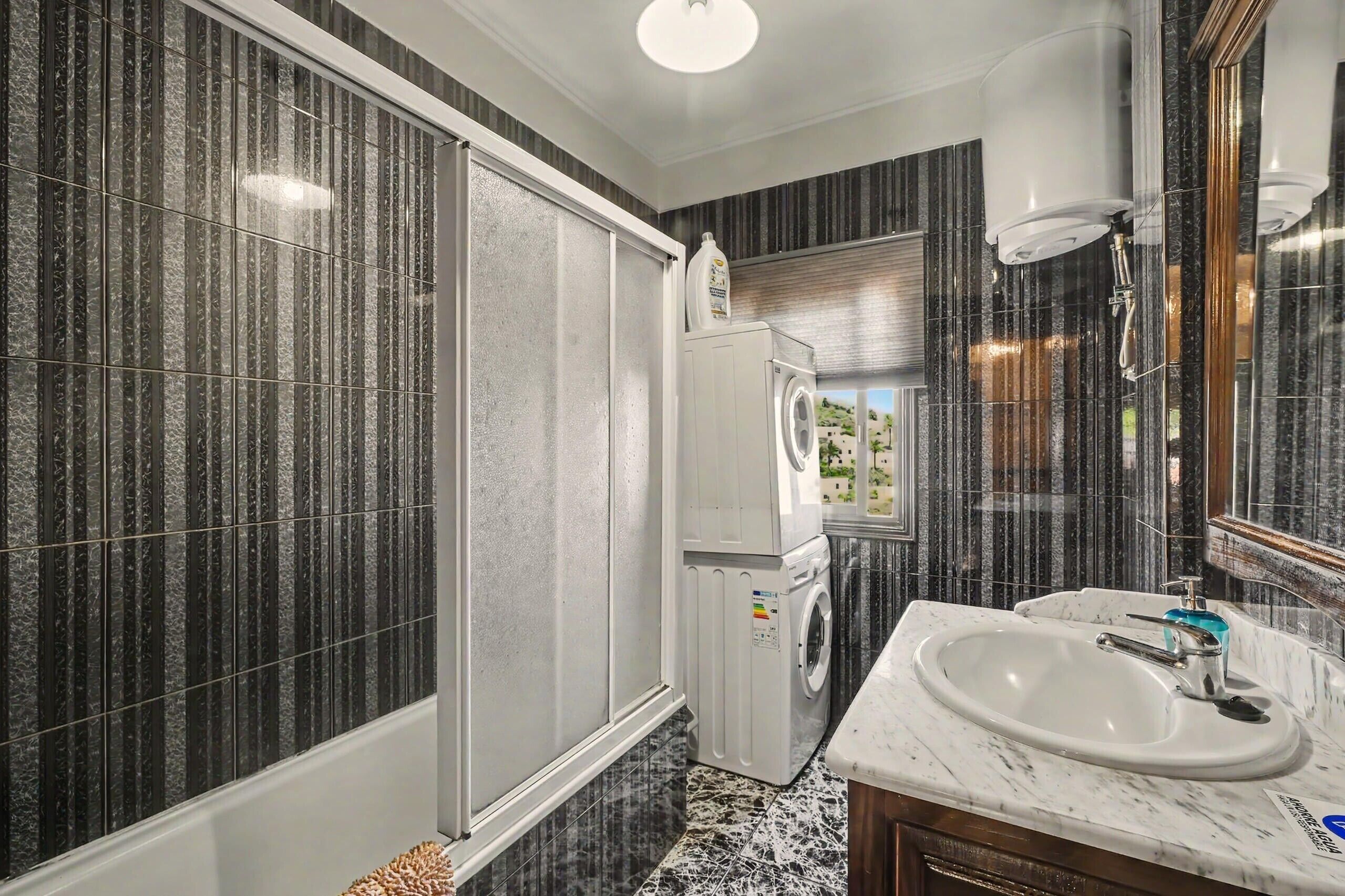 A bathroom with a large mirror, a sink, a washing machine, and a shower.