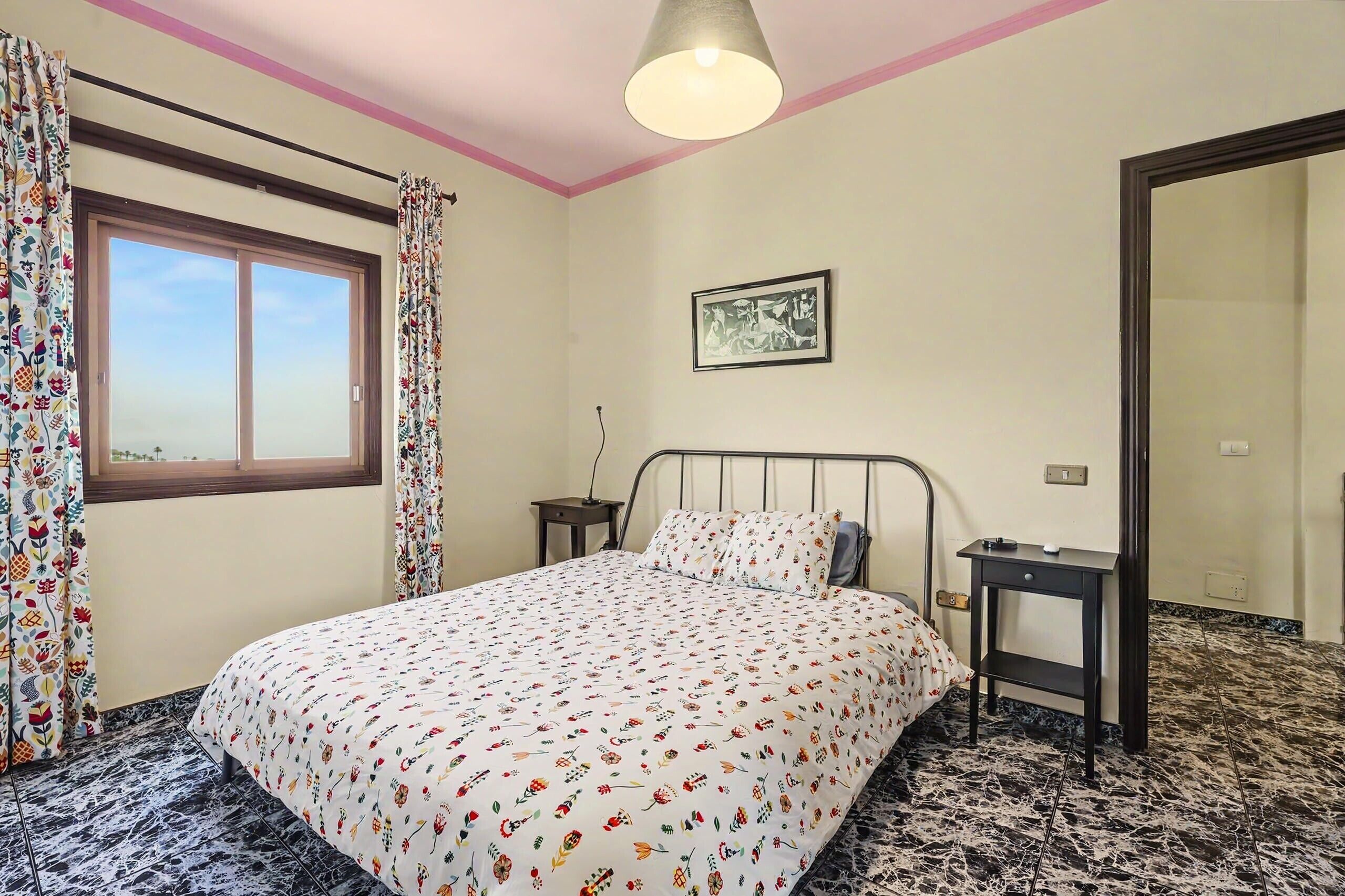 A bedroom with a bed, a bedside table, a nightstand, a window with curtains, a wall-mounted picture, and a door.
