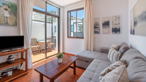 A living room with a sofa, a coffee table, a TV on a stand, and a balcony with a chair and table. - Casa Perla – Terrace & Pool in the Heart of Playa de Santiago (Playa de Santiago)
