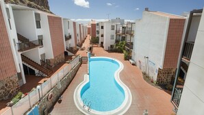 A swimming pool surrounded by multi-story buildings with balconies, a clear sky, and a rocky cliff in the background. - Casa Perla – Terrace & Pool in the Heart of Playa de Santiago (Playa de Santiago)