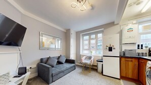 A modern living room with a grey sofa, a small white table, and a TV. There is a kitchen area with a fridge, microwave, and a washing machine. - Bright 1Bedroom Home-Brixton Tube 12min Away (Brixton)