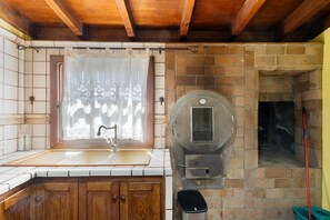 A kitchen with a brick oven, wooden cabinets, a sink, and a window with a lace curtain.