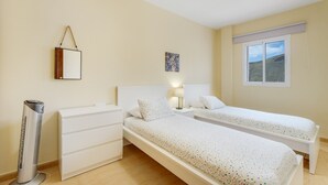 Un dormitorio con dos camas individuales, una mesita de noche con lámpara, un radiador y una ventana con vistas a una colina.