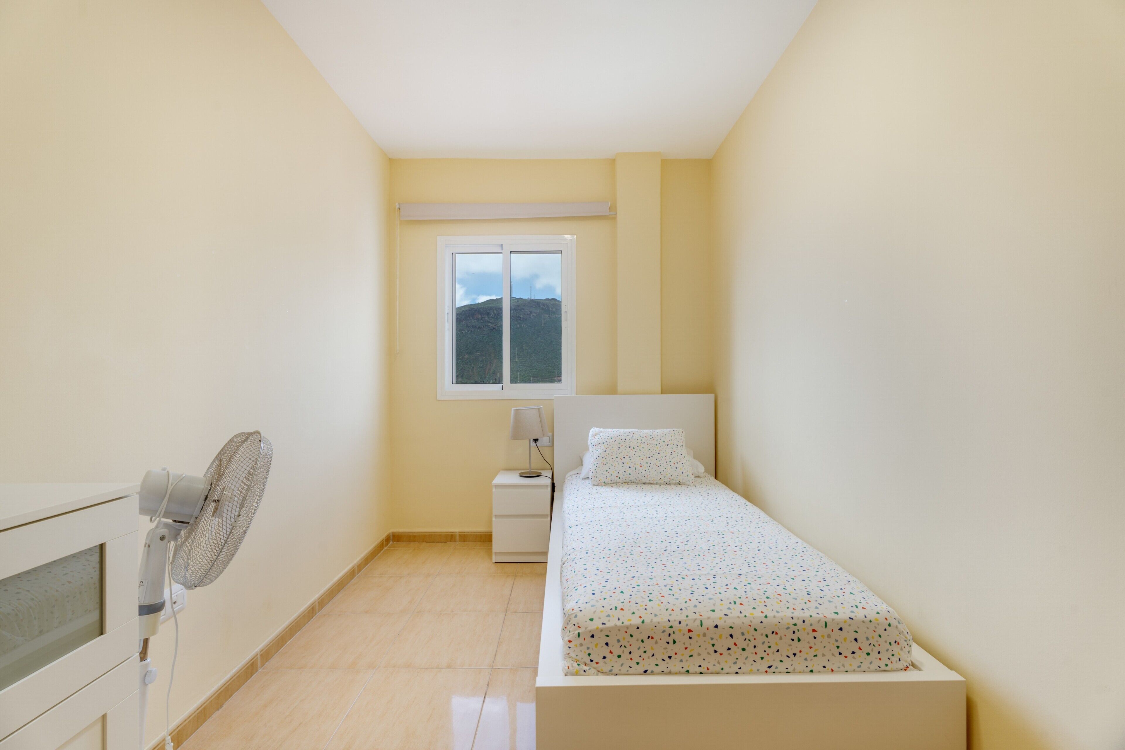 A small, single-bed room with a standing fan, a bedside table with a lamp, and a window with a view of a hill.