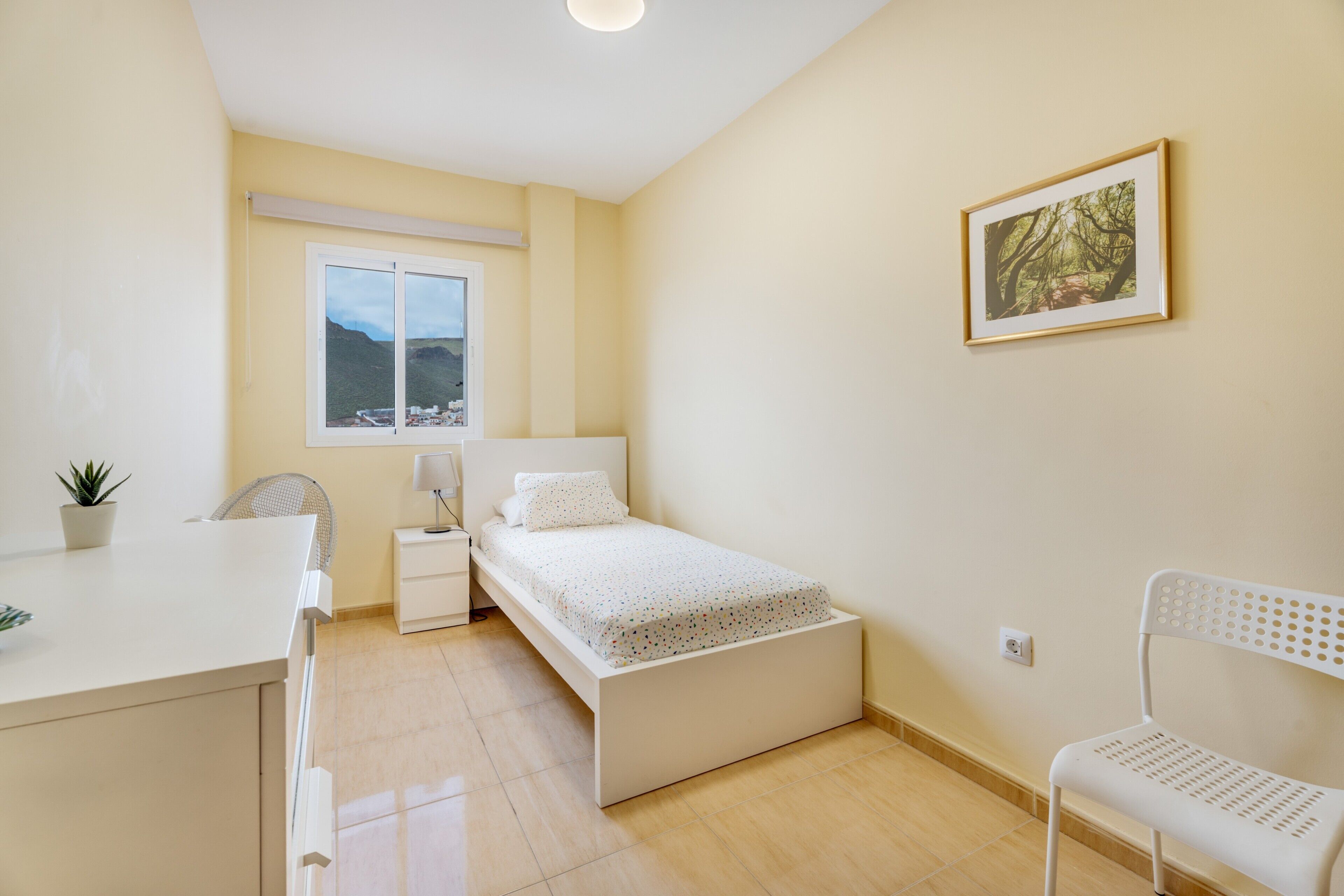 Een slaapkamer met een bed, nachtkastje, stoel, bureau, raam en een lijst met een afbeelding aan de muur.