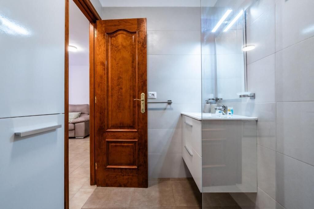 A bathroom with a wooden door, white tiled walls, a white sink, and a mirror.