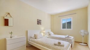 Un dormitorio con dos camas, una mesita de noche, un aparador, un espejo, una obra de arte en la pared, una ventana con vistas y un ventilador de pie.