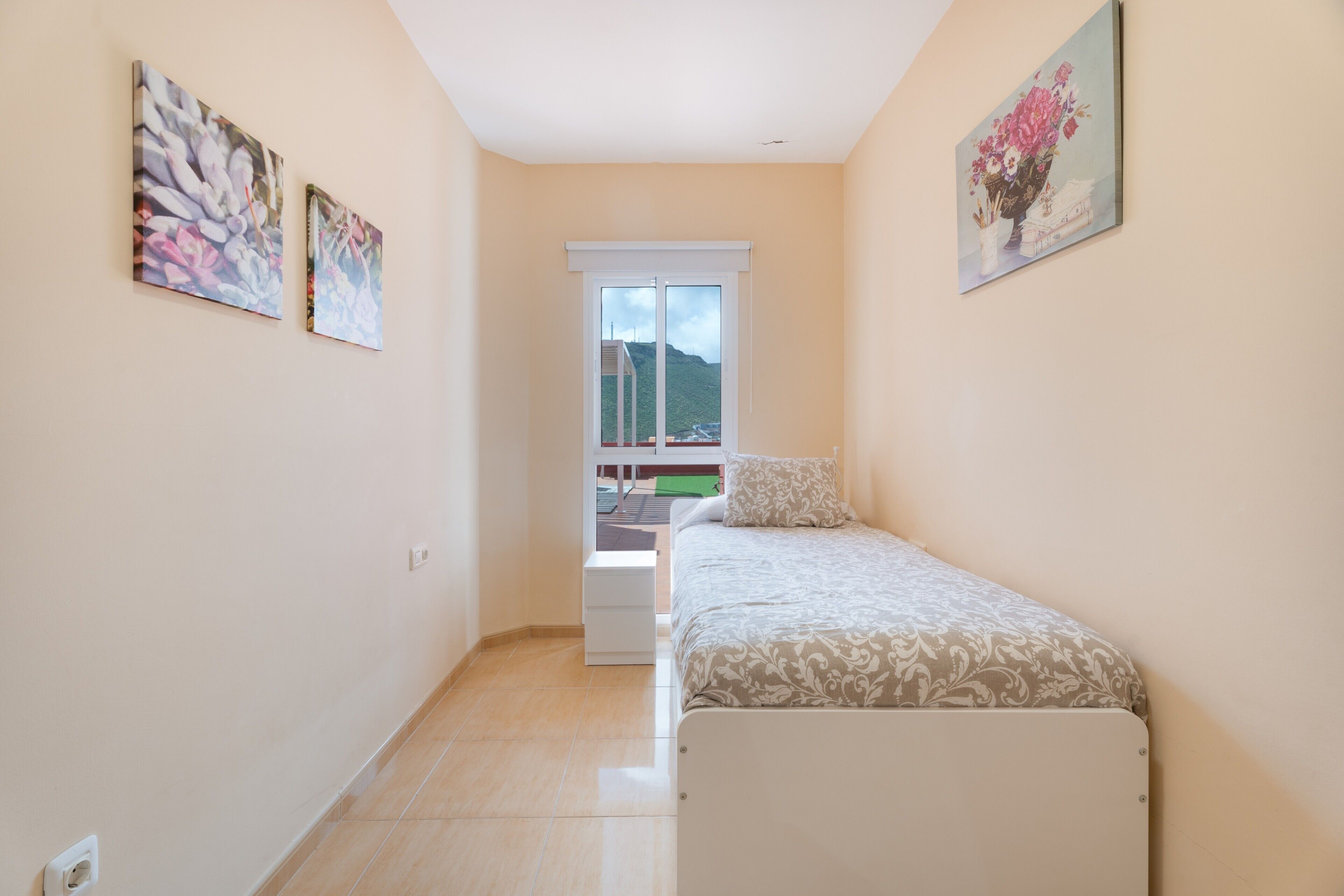A narrow room with a bed, a window, and two paintings on the wall.