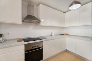 Una cocina moderna con armarios blancos, una campana extractora de acero inoxidable, un horno integrado y una encimera con un hervidor y una tabla de cortar.
