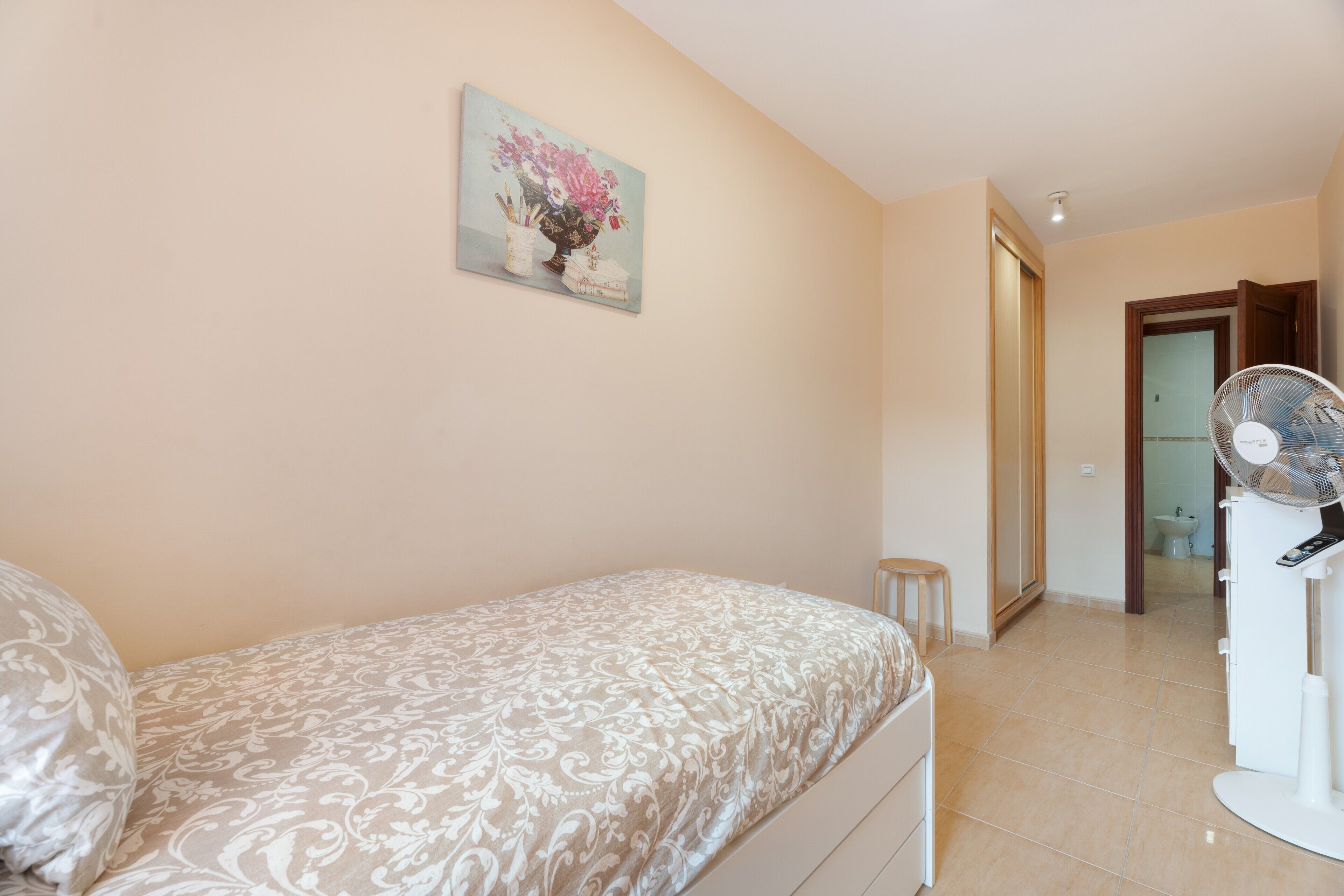Une chambre à coucher avec un lit, une table de chevet, un petit tabouret, un ventilateur et un tableau accroché au mur.
