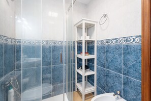 Un baño con azulejos de mármol azul, una cabina de ducha de vidrio, un estante blanco y un lavabo.