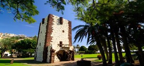 A historic tower with a brick facade and arched entrance, surrounded by a park with palm trees and a clear sky. - Casa Miramar 3 – Mountain Views & Central Location in San Sebastián (San Sebastian de La Gomera)