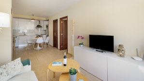 A modern living room with a sofa, a coffee table with a book and a bottle, a TV on a white cabinet, and a kitchen in the background. - Casa Miramar 2 – Comfort and Unbeatable Location in the Heart of San Sebastián (San Sebastian de La Gomera)