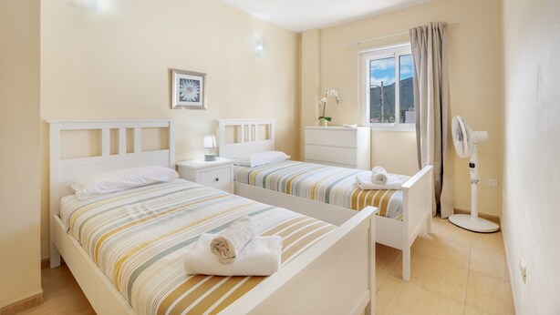 A bedroom with two beds, a window with curtains, a bedside table with a lamp, and a standing fan. - Casa Miramar 2 – Comfort and Unbeatable Location in the Heart of San Sebastián (San Sebastian de La Gomera)
