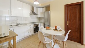 A modern kitchen with white cabinets, stainless steel appliances, a round dining table with chairs, and a wooden door. - Casa Miramar 2 – Comfort and Unbeatable Location in the Heart of San Sebastián (San Sebastian de La Gomera)