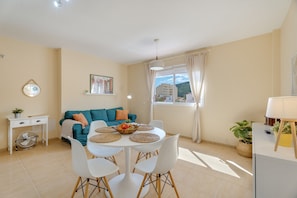 Un salon moderne avec un canapé de couleur turquoise, une table à manger avec des chaises blanches, un petit bureau blanc et une plante en pot.