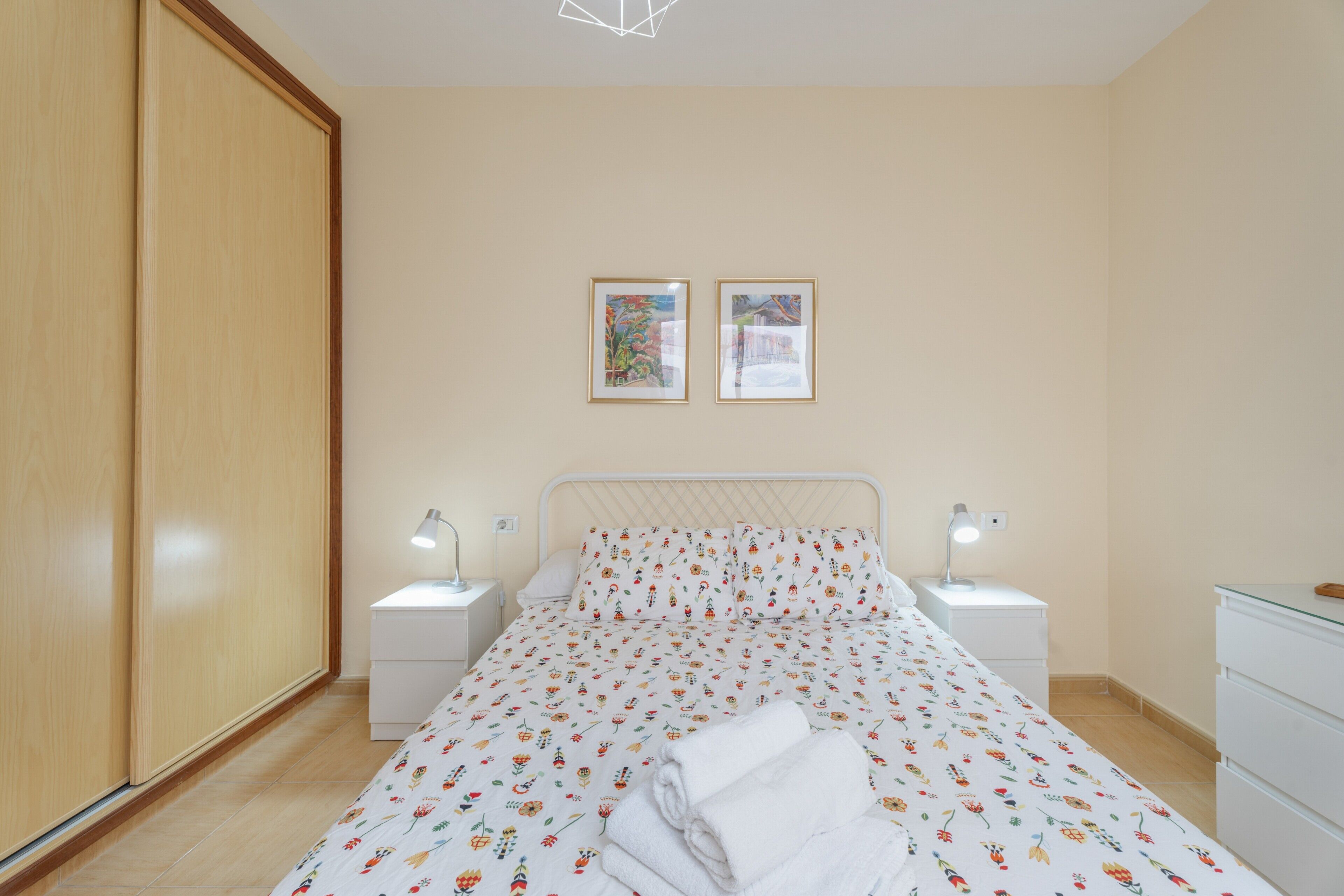 A bedroom with a bed, two bedside tables, a wardrobe, and two framed pictures on the wall.