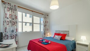 Een slaapkamer met een bed, nachtkastjes, een bureau, een ventilator en een raam met gordijnen.