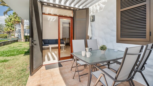 Una terrazza aperta con un tavolo in vetro, sedie e una porta scorrevole che dà su una zona salotto interna.
